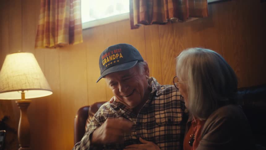 An elderly couple affectionately looks through a photo album, sharing a warm embrace while reminiscing, with a close-up on the man's 'World's Best Grandpa' hat