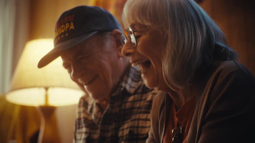 A heartwarming moment as an elderly couple laughs together while reminiscing and looking through a photo album in their living room