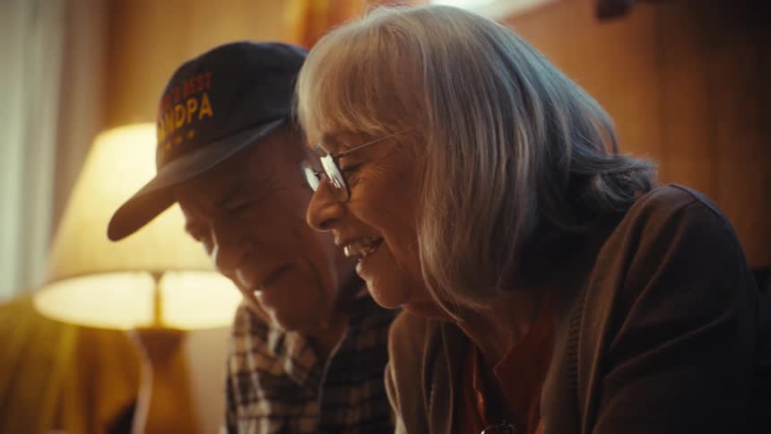 An elderly couple sits together on a couch in their warm living room, smiling as they reminisce over shared memories