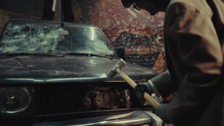 An angry man in a suit and protective face shield smashes a car's hood with a sledgehammer in a gritty, graffiti-covered garage in slow motion