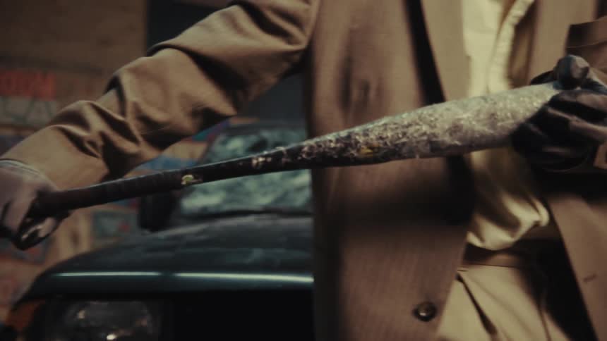A menacing figure in a suit and gloves holds a dirty baseball bat in a gritty urban garage