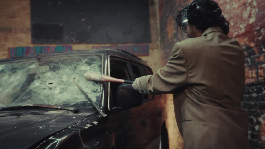 Slow motion shot of a man in a business suit and protective gear destroying a car with a baseball bat in a rage room