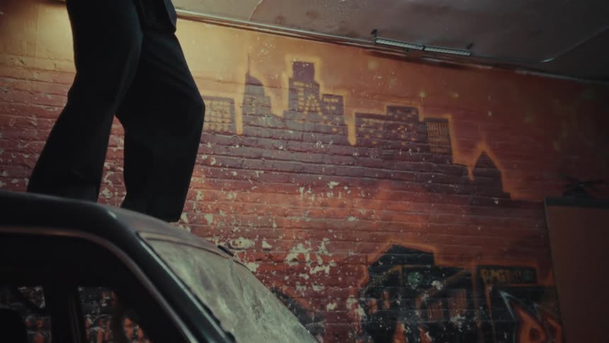 An angry man in a suit and protective helmet smashes a car's windshield with a sledgehammer in slow motion inside a gritty garage