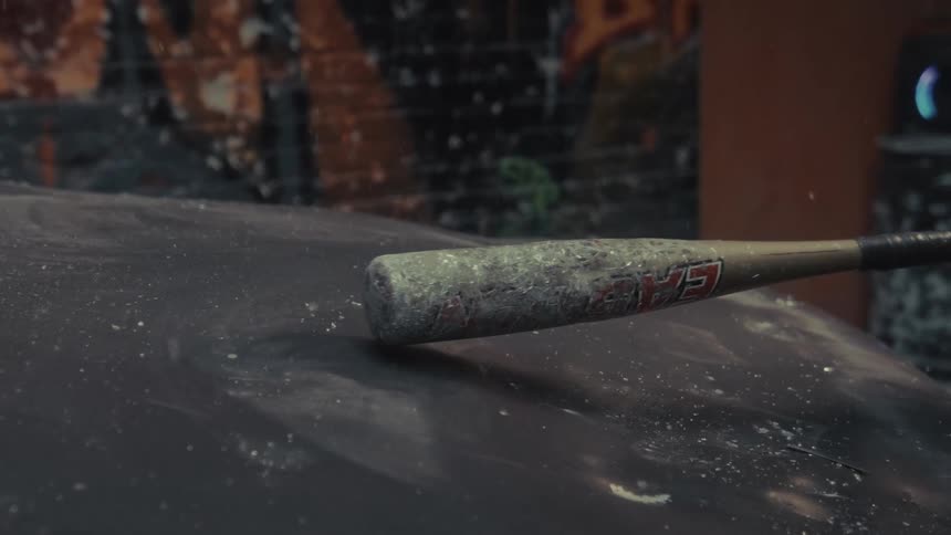 A baseball bat covered in dust and debris hits the hood of a car with force, captured in super slow motion