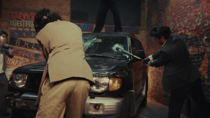 Business people in suits and protective gear take out their frustrations by smashing a car with sledgehammers and bats in slow motion