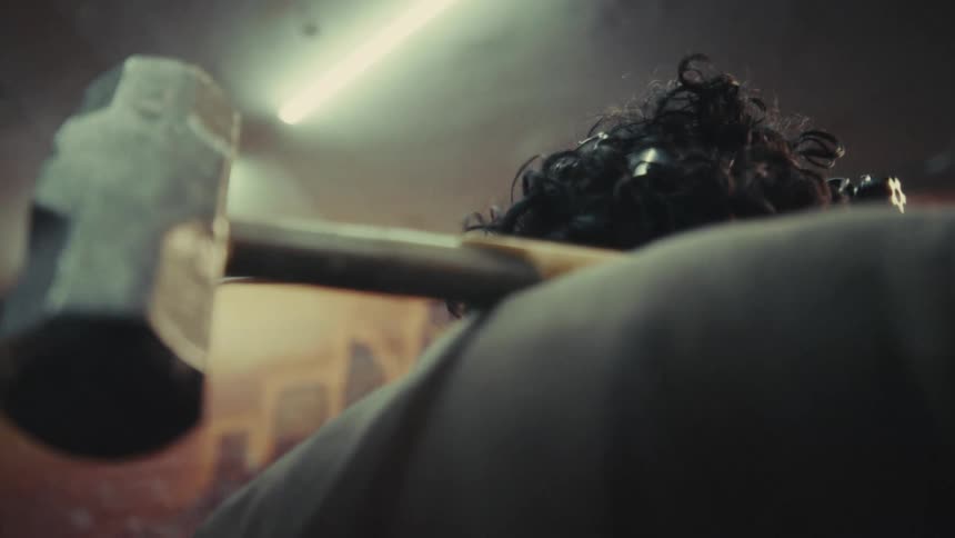 A gritty, cinematic over-the-shoulder shot reveals a person with a sledgehammer in a dimly lit, industrial space