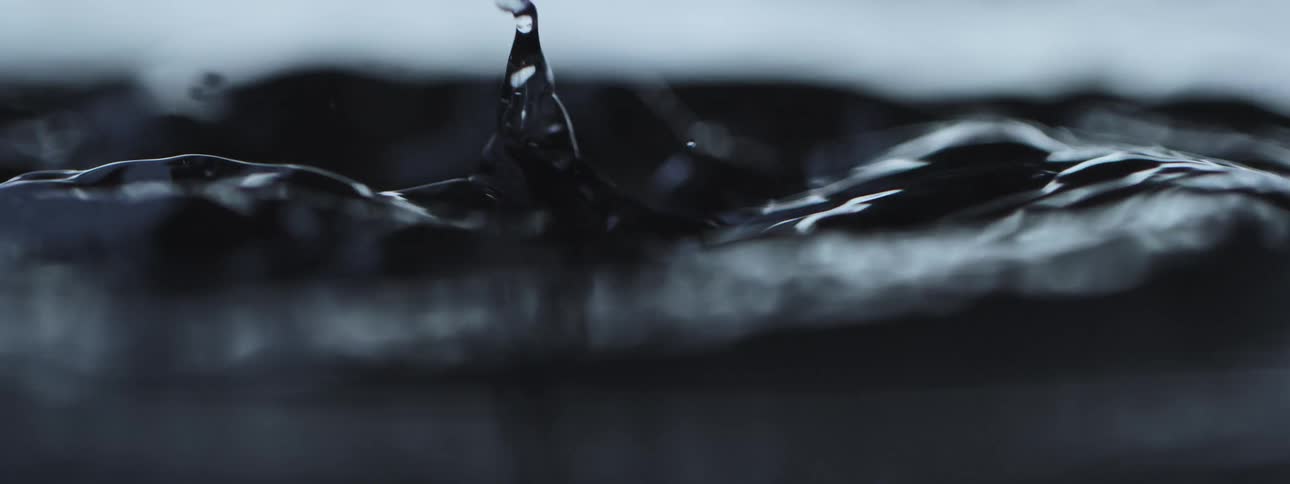 An extreme close-up of a single water droplet falling into dark water in slow motion, creating ripples