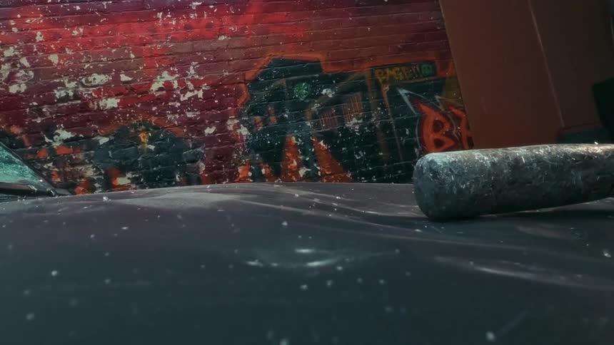 A metal baseball bat smashes the dusty hood of a car in a gritty urban alleyway in slow motion