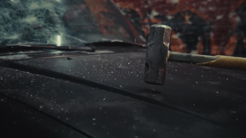 A sledgehammer crashes down onto the hood of a dusty black car in dramatic slow motion, sending debris flying
