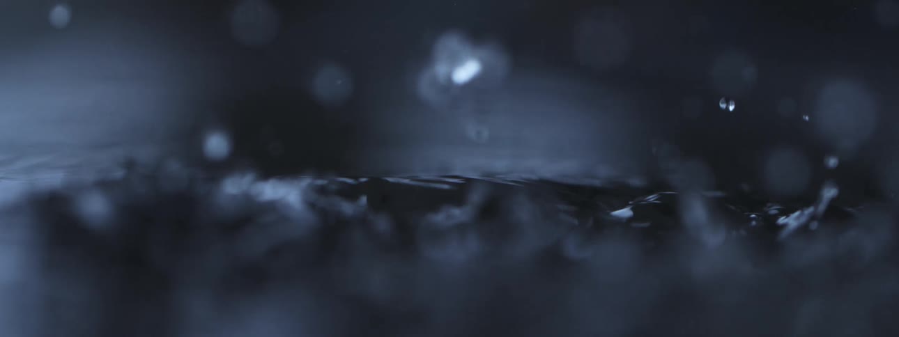 A dramatic and mesmerizing slow-motion macro shot of raindrops splashing onto a dark surface of water, creating ripples and bokeh