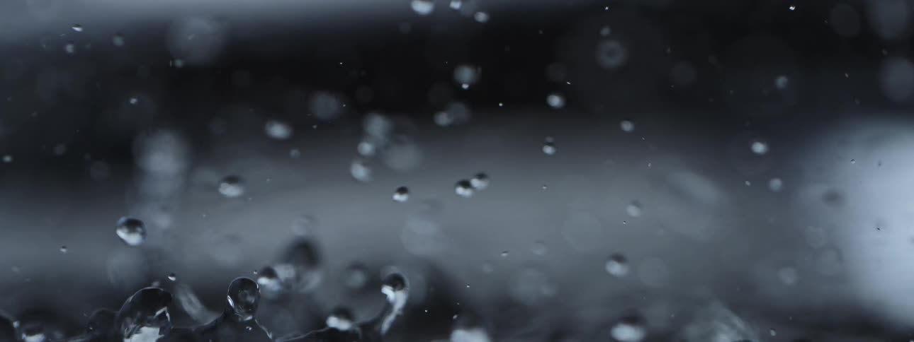 A macro slow motion shot shows water droplets splashing and bouncing against a dark, moody background