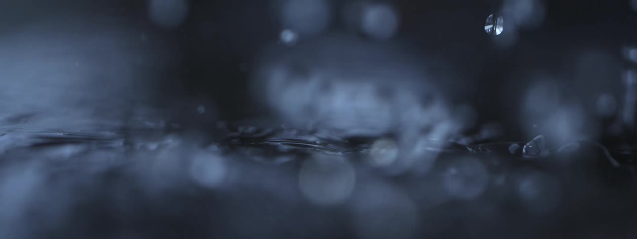 A macro slow-motion view of raindrops splashing into dark water, creating beautiful ripples and bokeh