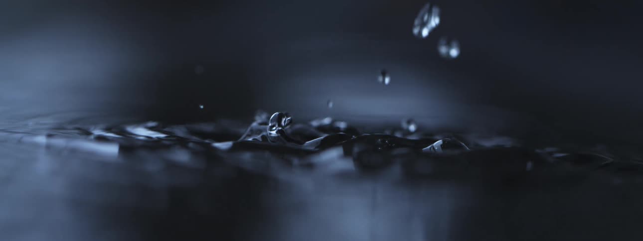 A dramatic super slow-motion close-up of water droplets splashing into dark, still water, creating intricate patterns and ripples