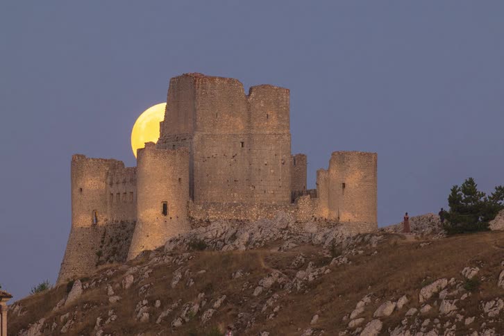 A stunning timelapse captures the huge, golden full moon rising behind the ancient Rocca Calascio castle as people gather to watch the celestial event at twilight