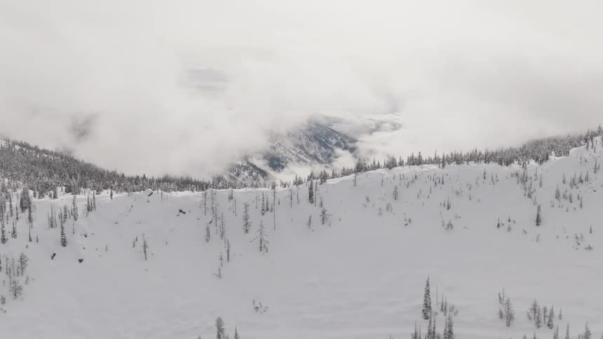 An aerial view pans over a vast, snow-covered mountain landscape with sparse trees under a thick blanket of clouds