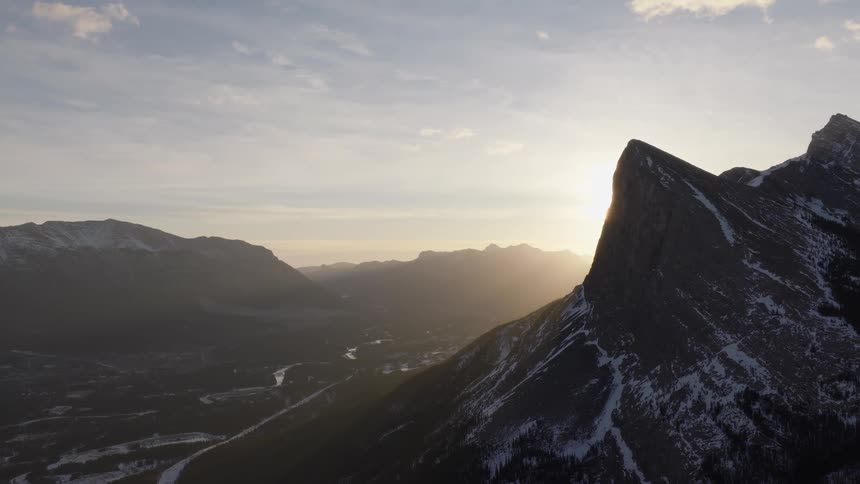 A stunning sunrise emerges from behind a jagged, snow-capped mountain, casting a beautiful flare over the landscape