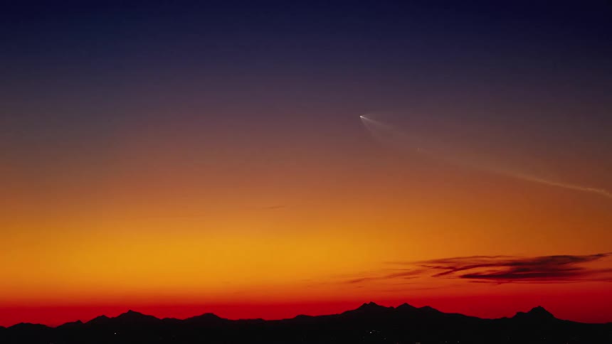 A rocket creates a visual phenomenon across the vibrant, colorful sky just after sunset, flying over a silhouetted mountain range