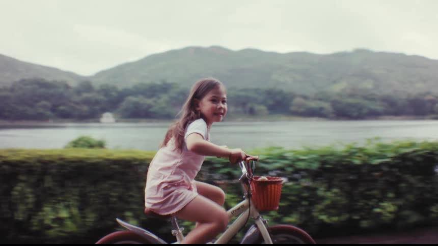 A happy young girl smiles while riding her bike with training wheels along a scenic lakeside path in a park
