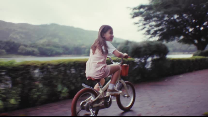A happy young girl smiles while riding her bike with training wheels along a scenic lakeside path in a park