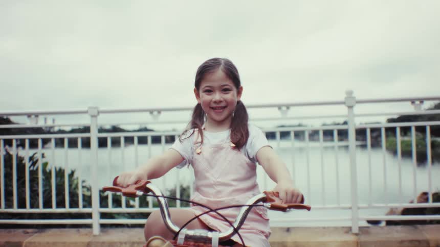 A cheerful young girl with pigtails sits on her bicycle and smiles happily at the camera in slow motion