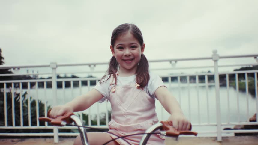 A cheerful young girl with pigtails sits on her bicycle and smiles happily at the camera in slow motion