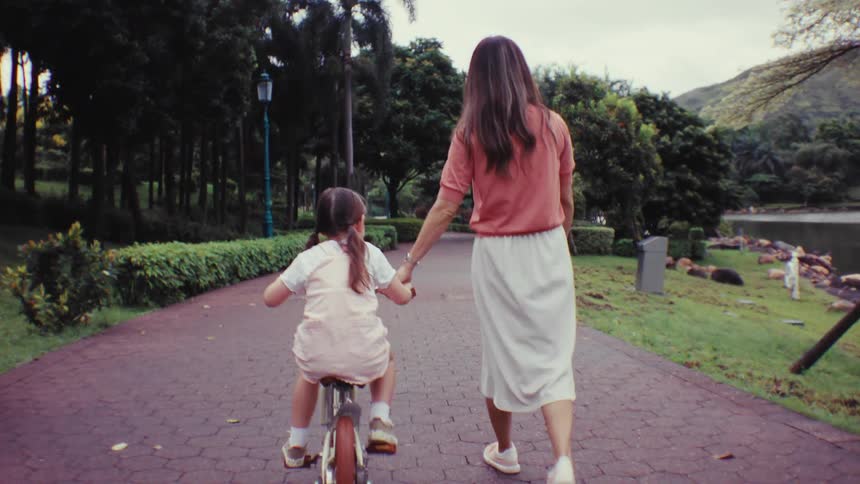 A mother holds her daughter's hand and helps her ride a bike for the first time on a path through a lush green park