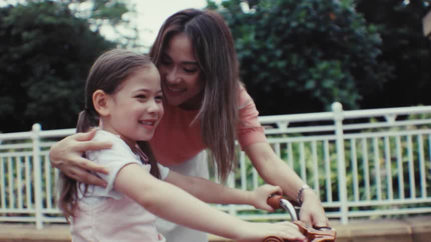 A loving mother encourages her young daughter who smiles brightly while learning to ride her bicycle in the park