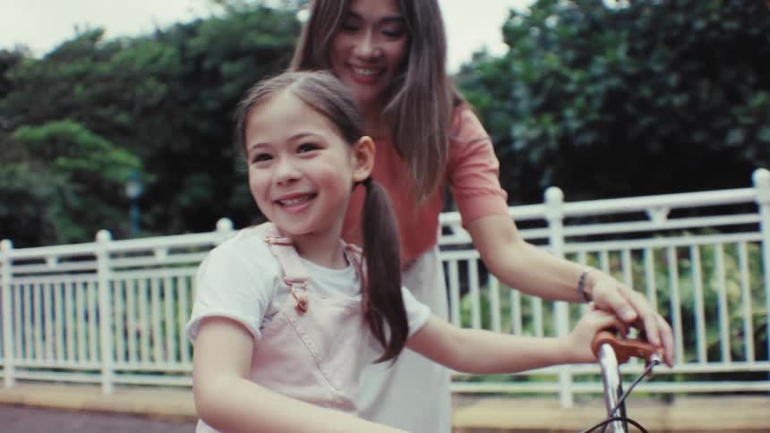 A loving mother encourages her young daughter who smiles brightly while learning to ride her bicycle in the park