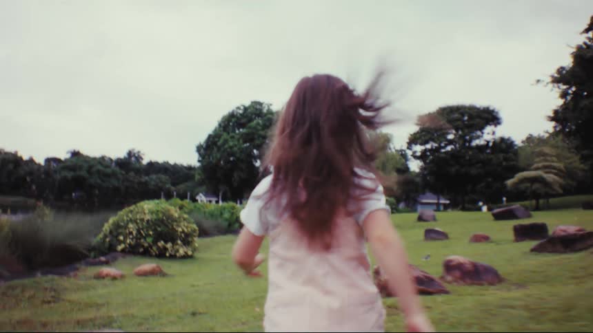 A carefree young girl with long flowing hair runs through a green park in a dream-like slow motion shot