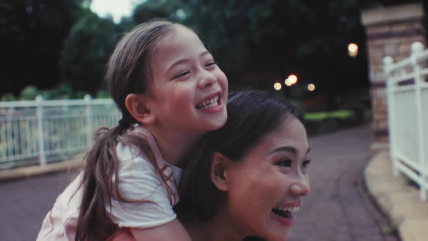 A happy little girl laughs joyfully while getting a piggyback ride from her loving mother in a park