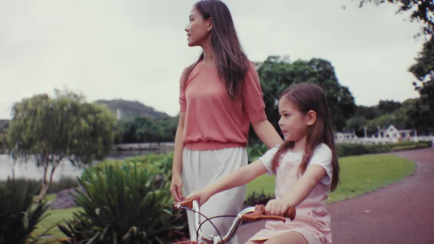 A loving mother teaches her daughter to ride a bicycle in a beautiful park, pointing the way forward as the girl happily starts to pedal