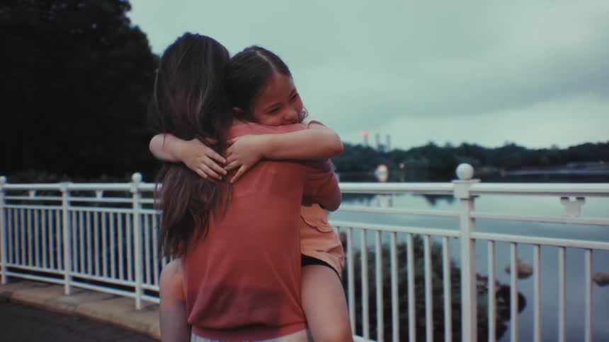 A mother holds her young daughter in her arms, and the smiling girl gives her a tight, loving hug in slow motion by a lake at dusk