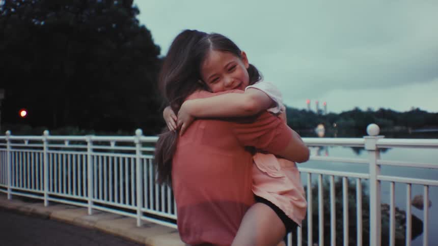 A mother holds her young daughter in her arms, and the smiling girl gives her a tight, loving hug in slow motion by a lake at dusk
