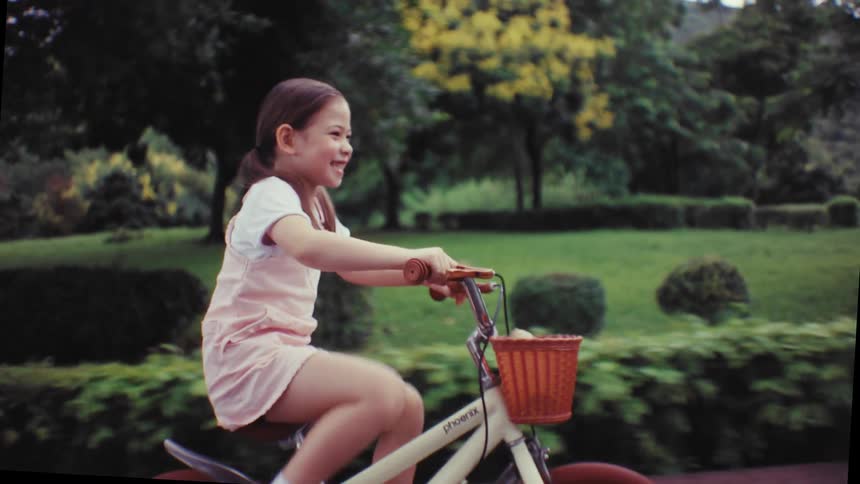 A joyful little girl with a big smile rides her bicycle in slow motion along a path in a beautiful green park