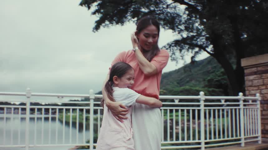 A happy little girl runs across a park bridge in slow motion and into her smiling mother's loving embrace