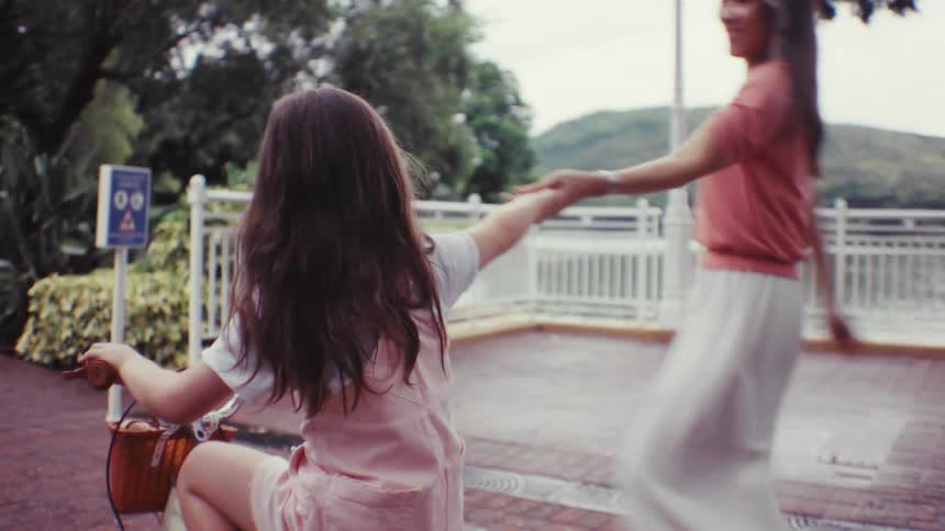 A joyful young girl riding her bicycle on a park path looks back and holds hands with her mother walking alongside her