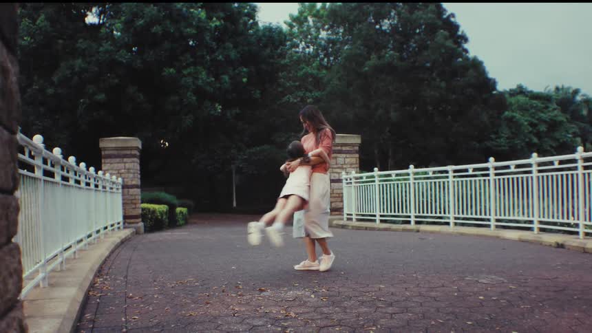 A loving mother playfully spins her happy young daughter around on a paved path in a beautiful green park