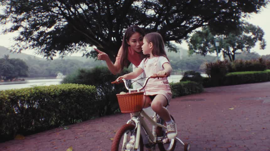 A mother and her young daughter pause on a park path while bike riding to point at something interesting by the lake
