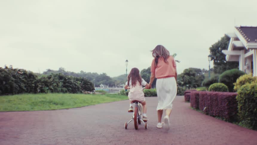 A caring mother teaches her happy young daughter how to ride a bicycle with training wheels along a scenic park path