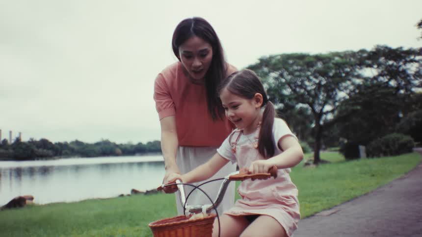 A mother teaches her happy daughter to ride a bike in a lakeside park, then watches her ride off on her own