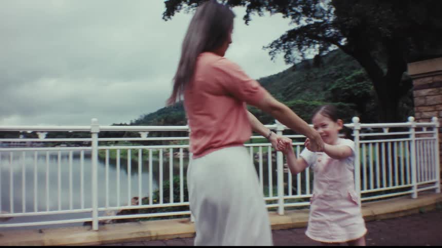 A mother and her young daughter joyfully hold hands and spin in a circle together in slow motion while in a park by a lake