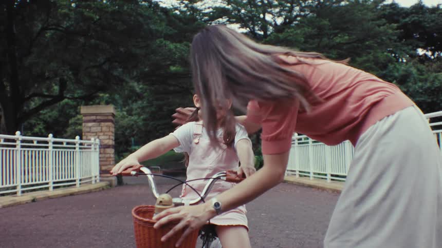 A loving mother encourages her happy daughter learning to ride her first bicycle in a beautiful park, sharing a kiss in slow motion