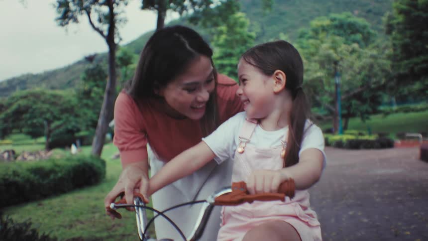 A caring mother teaches her smiling daughter how to ride a bicycle for the first time in a beautiful park in slow motion