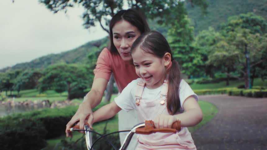 A caring mother teaches her smiling daughter how to ride a bicycle for the first time in a beautiful park in slow motion