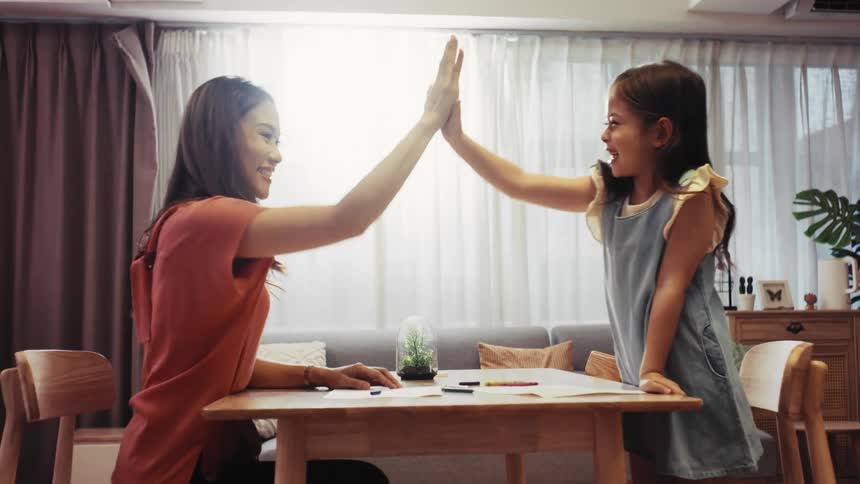 A loving mother and daughter share a joyful high-five in slow motion over a drawing table in their sunlit home