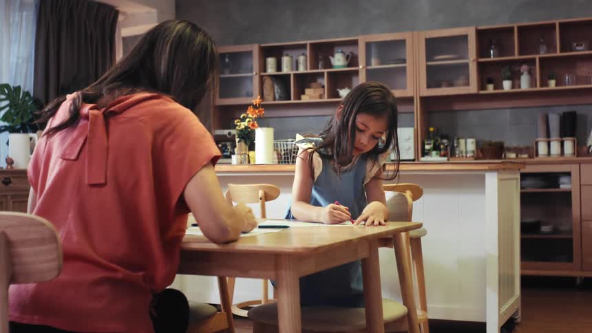 A young girl and her mother sit side-by-side at a kitchen table, enjoying a creative afternoon coloring together