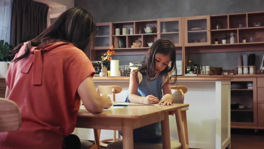 A young girl and her mother sit side-by-side at a kitchen table, enjoying a creative afternoon coloring together