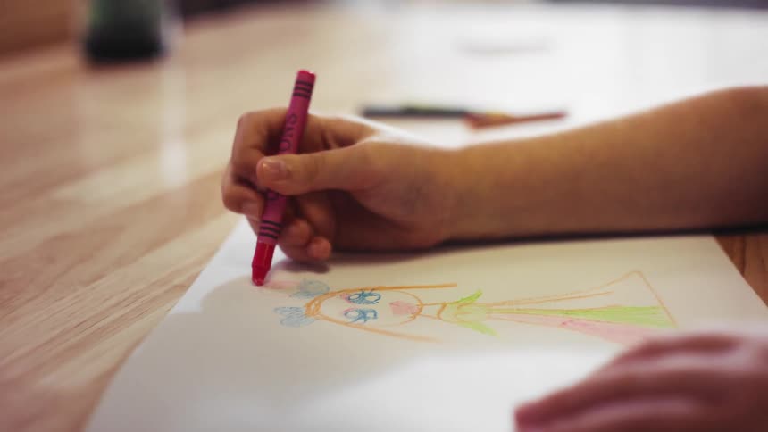 A close up of a child's hands coloring in a drawing of a person with a pink crayon on a wooden surface