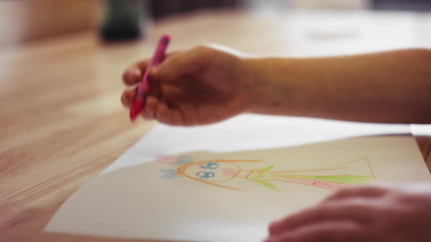 A close up of a child's hands coloring in a drawing of a person with a pink crayon on a wooden surface