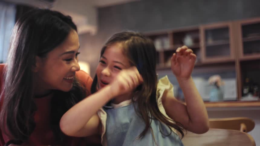 A happy mother and her playful daughter laugh and smile while drawing together with crayons at home